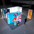 A vibrant exhibition stand featuring illuminated displays with stylish models wearing modern sunglasses and bright neon colors. The setup includes a bold blue panel with three fashionable women, a smaller display with a model in sunglasses, and a tall orange panel showcasing a chic handbag.